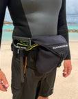 Person wearing a wetsuit and boardshorts with a waist bag on a beach.
